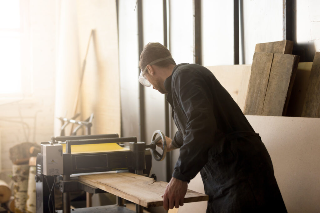 Back view of young carpenter or craftsman wearing working clothes and protective eyewear woodworking in small workshop interior, operating stationary power planer, processing trimming wooden plank