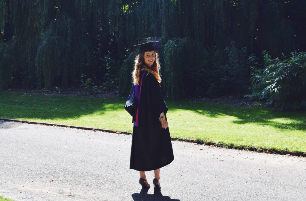 A picture of me on my graduation day, in front of greenery, wearing black and purple graduation robe.