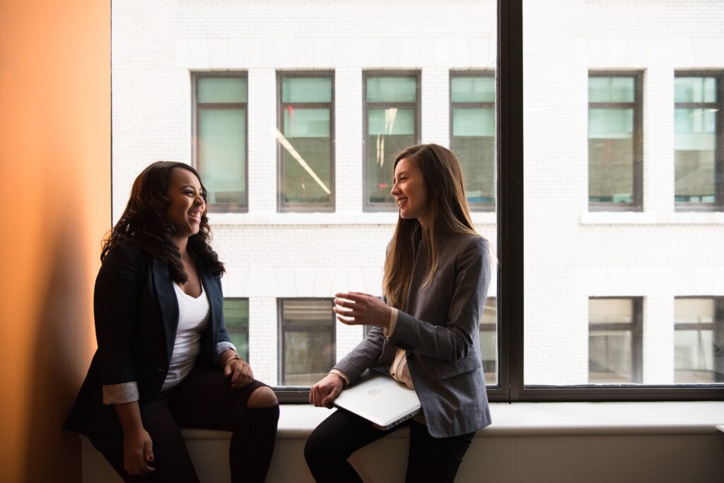 Two smiling women are dressed in smart clothing and sitting on a large, bright window sill. 