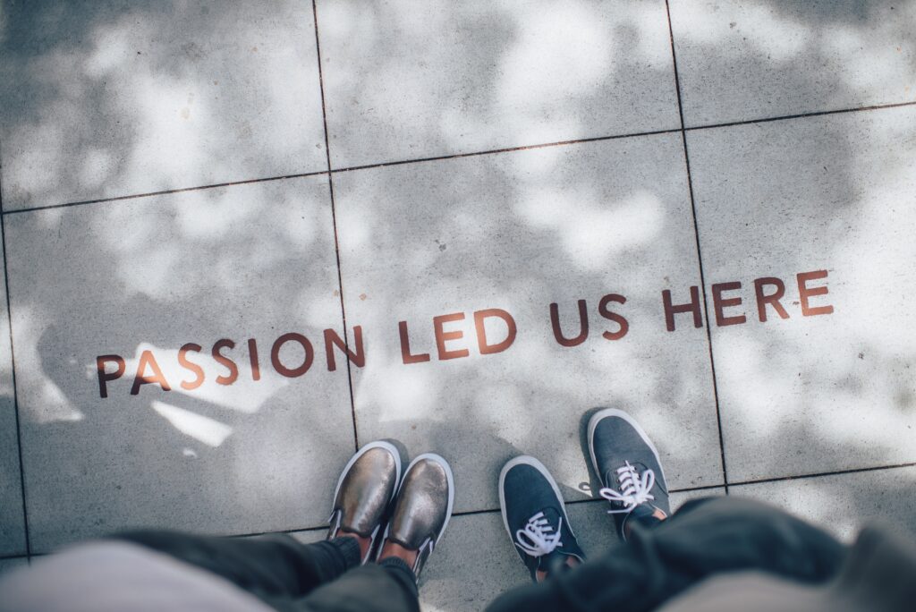 Two people who are only visible by their feet. They stand above writing on the pavement which says: Passion led us here