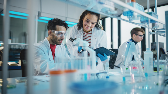 Portrait of Healthcare Science Practitioners Using Microscope, Digital Tablet, Doing Sample Analysis, Talking. Diverse Team of Specialists working in an Advanced Lab.